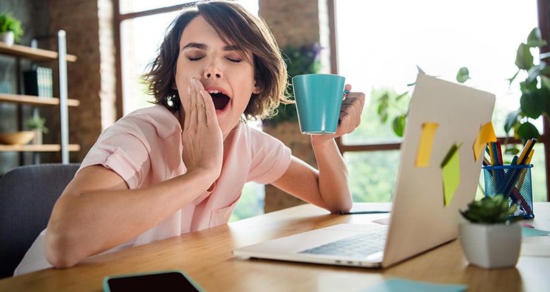 woman yawning, holding coffee cup sitting at computer