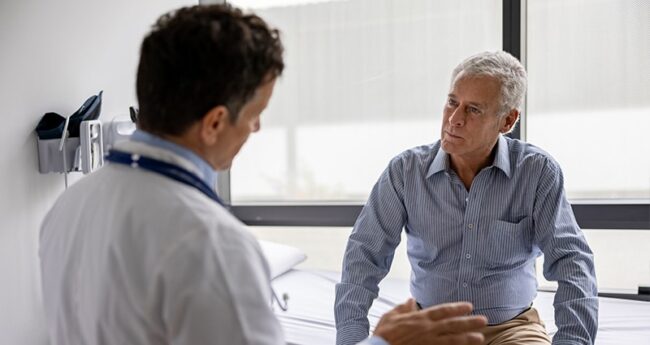 MensHealthScreening_Featured A medical professional wearing a white coat is facing away from us. He's explaining something to a man wearing a blue shirt and sitting on an exam table. The man listens carefully.