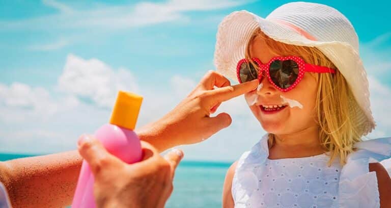 A little girl wearing a hat and heart-shaped sunglasses smiles as an adult dabs sunscreen on her nose. She's standing in front of a bright blue expanse of water.