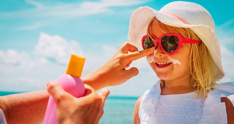 A little girl wearing a hat and heart-shaped sunglasses smiles as an adult dabs sunscreen on her nose. She's standing in front of a bright blue expanse of water.