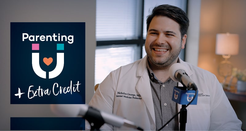 A man wearing a medical coat sits next to a screen that reads "Parenting U Extra Credit." He is smiling. There's a microphone in front of him.