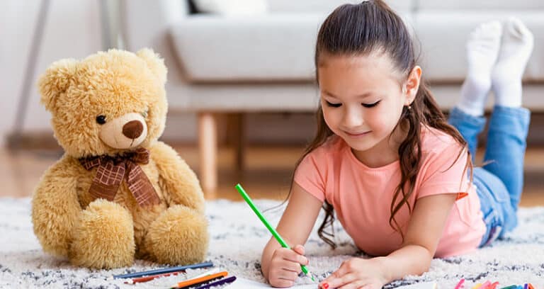 girl coloring on floor with teddy bear