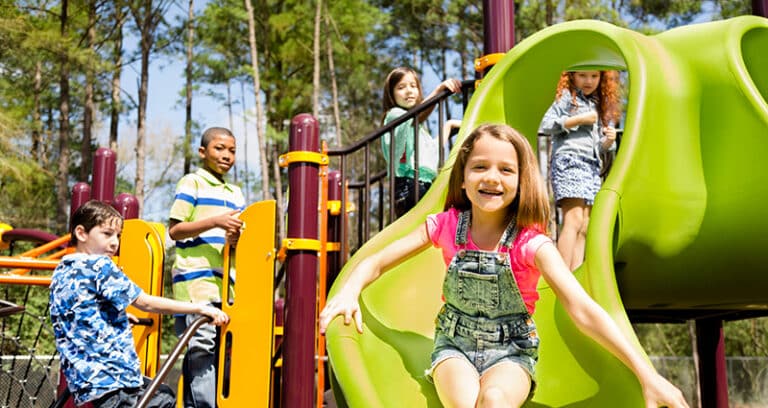Children play on a playground. Nearest the viewer, a little girl emerges from a covered, curving green slide.