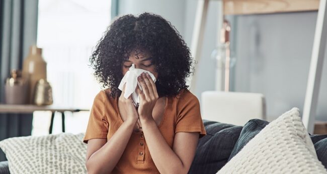 FluFAQ_Featured A woman sits on a sofa with pillows. She is blowing her nose.