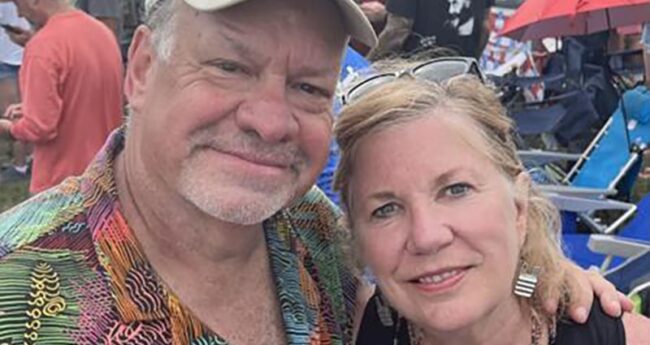 A man and woman smile at the camera. The man is wearing a cap and a colorful shirt. The woman's head is leaned close to him.