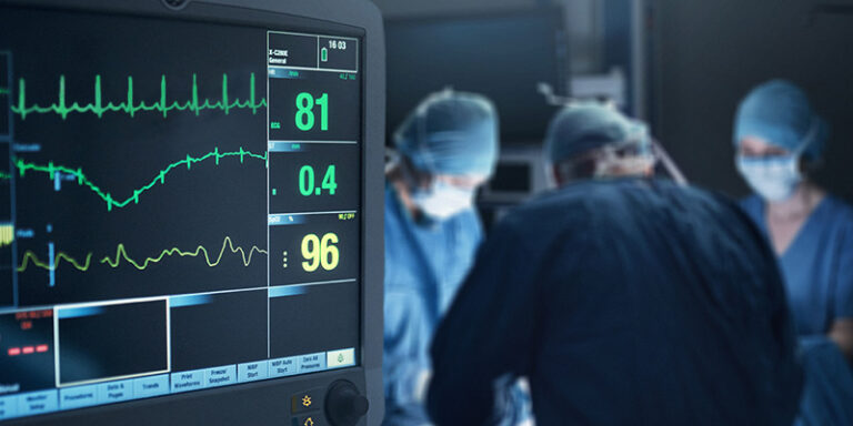 A heart monitor is in the foreground while surgeons are performing surgery in the background
