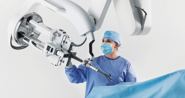 A surgeon wearing blue scrubs and a mask working with a surgical robot